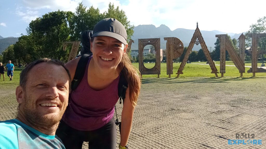 Kamila and Nick smiling in front a big wooden sign Zakopane. Sunny, summer weather. Giewont (Tatras) in the background.